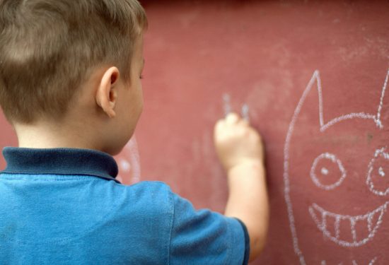 A young boy draws with white chalk on a red wall, creating a smiling face with pointy ears.