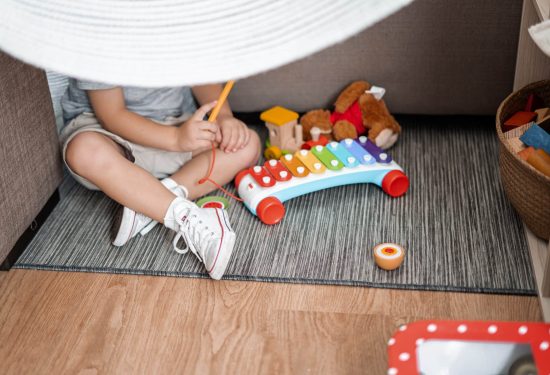 Child sitting on rug playing with xylophone, surrounded by toys and wearing white sneakers under a white canopy.