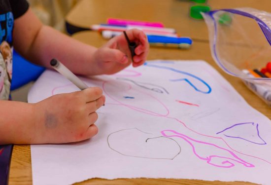 Child drawing colorful shapes on white paper with markers at a table.