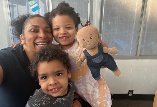 Smiling adult with two young children and a doll, posing for a cheerful selfie indoors.