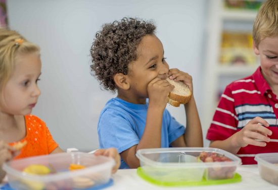 Three young children sit at a table eating lunch from plastic containers, smiling and enjoying their food.