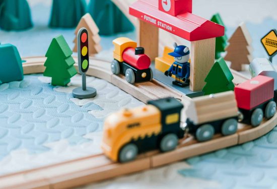 Wooden toy trains on tracks near a station, traffic light, trees, and a toy figure in a blue uniform.