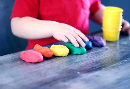 A child in a red shirt touches colorful playdough arranged in a row on a table.