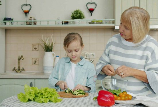 A young girl slices cucumbers while an older woman prepares salad in a kitchen with fresh vegetables.